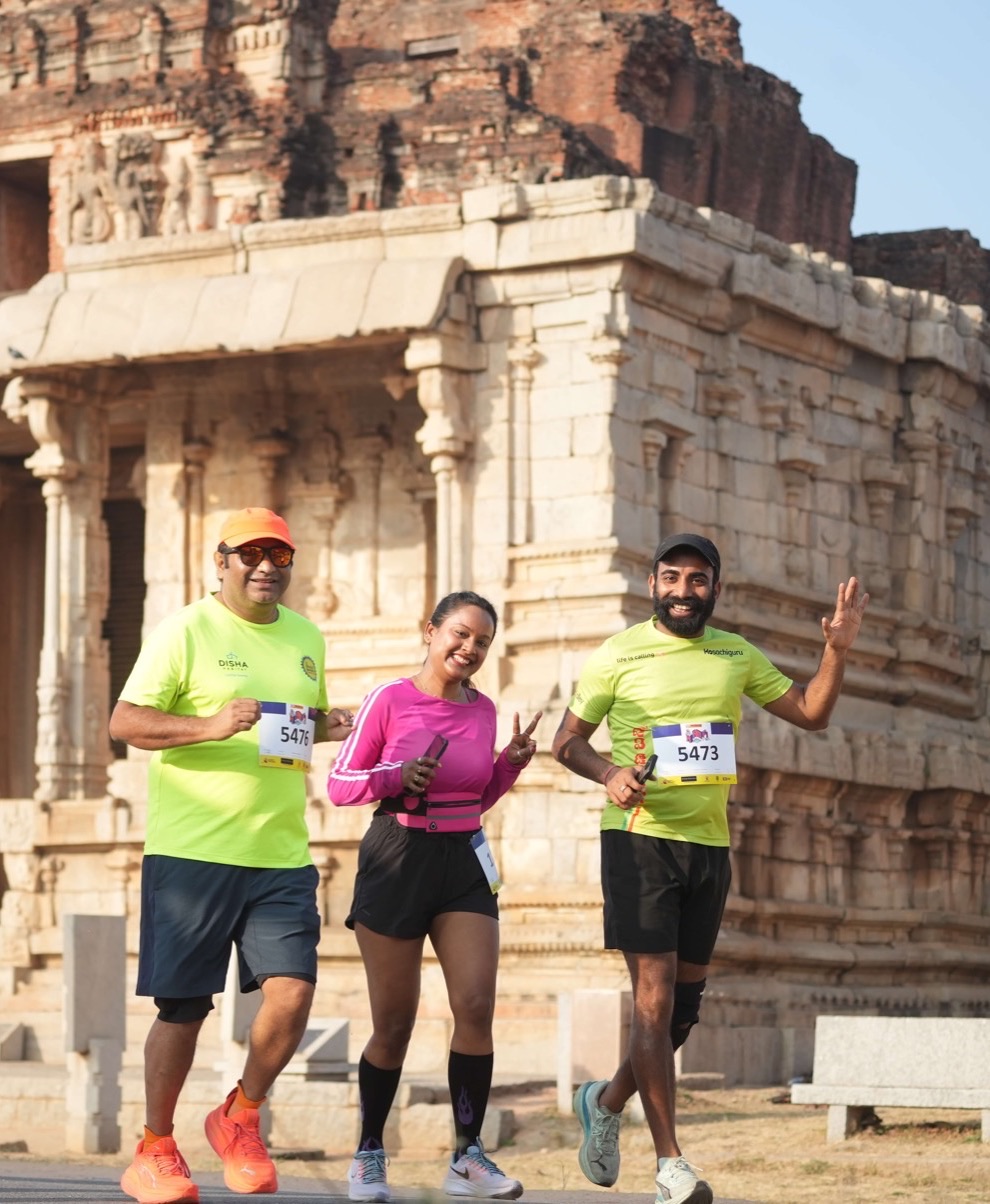 Three runners with bibs wave as they pass a Vijayanagara temple at GHR Hampi